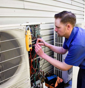 A technician in a blue shirt works on the wiring of an outdoor air conditioning unit mounted next to a house.