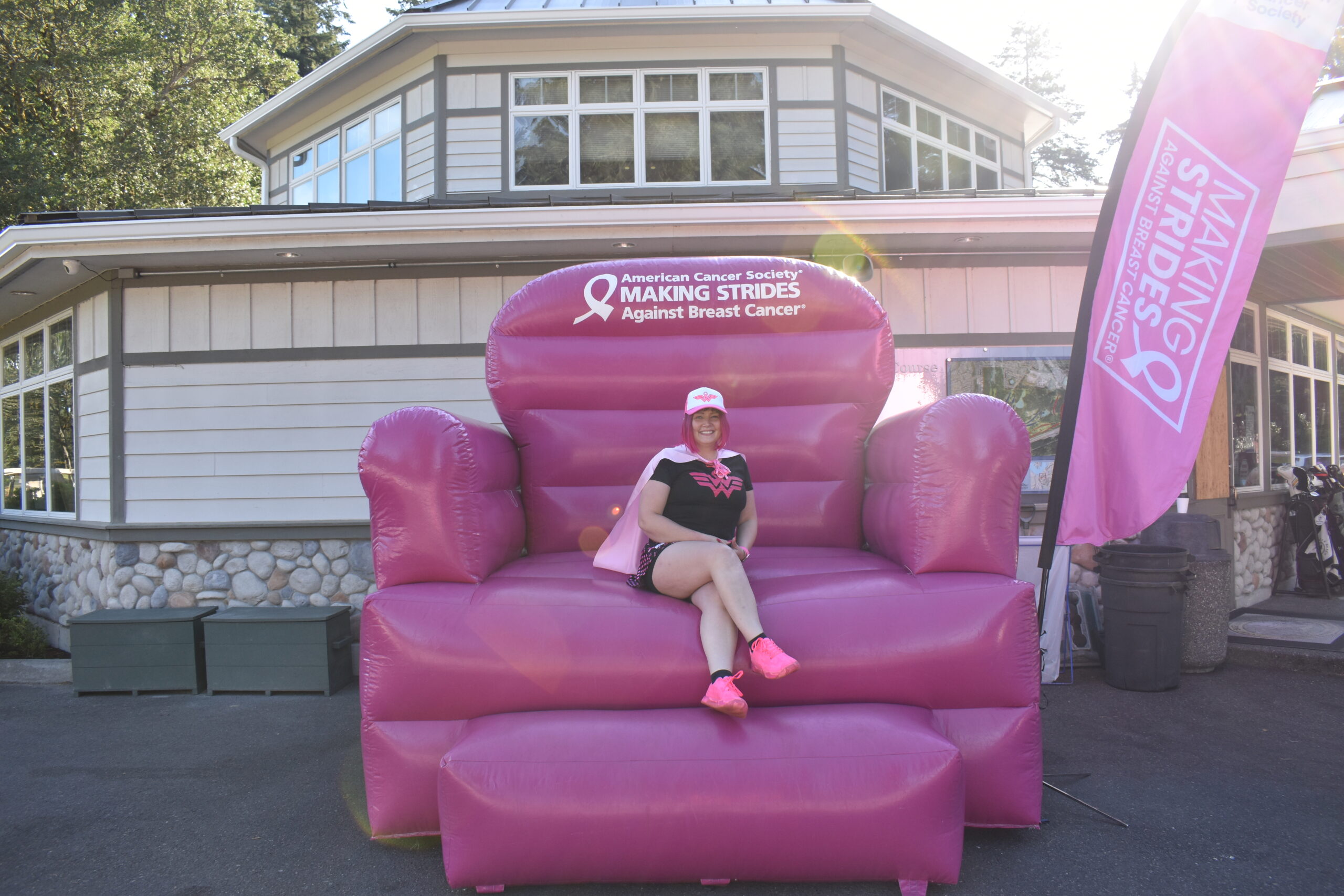 A woman in pink sits on a giant inflatable pink chair with "Making Strides Against Breast Cancer" written on it, next to a matching flag, promoting Golf Registration in front of a building.