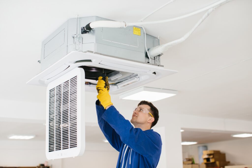A technician in blue overalls and yellow gloves is installing or repairing a ceiling-mounted air conditioning unit in a white room.