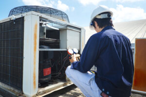 A technician wearing a hard hat uses a gauge to inspect and service a rooftop air conditioning unit under a clear sky.