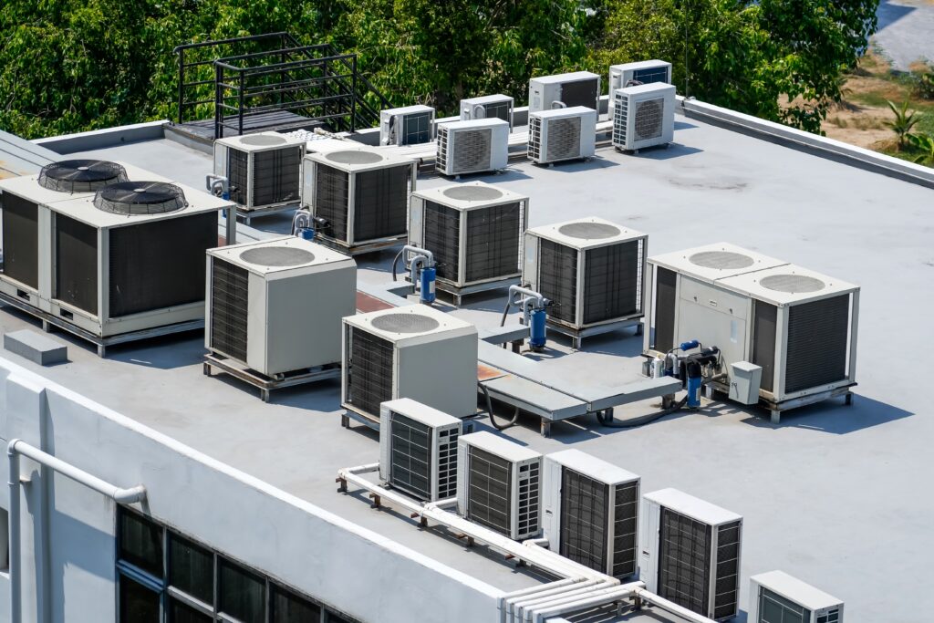 Multiple large air conditioning units and ventilation systems are installed on a flat building rooftop, surrounded by trees and outdoor scenery.