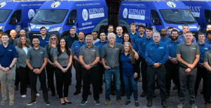 A group of employees poses in front of several blue Kiefmann Services vans parked side by side.