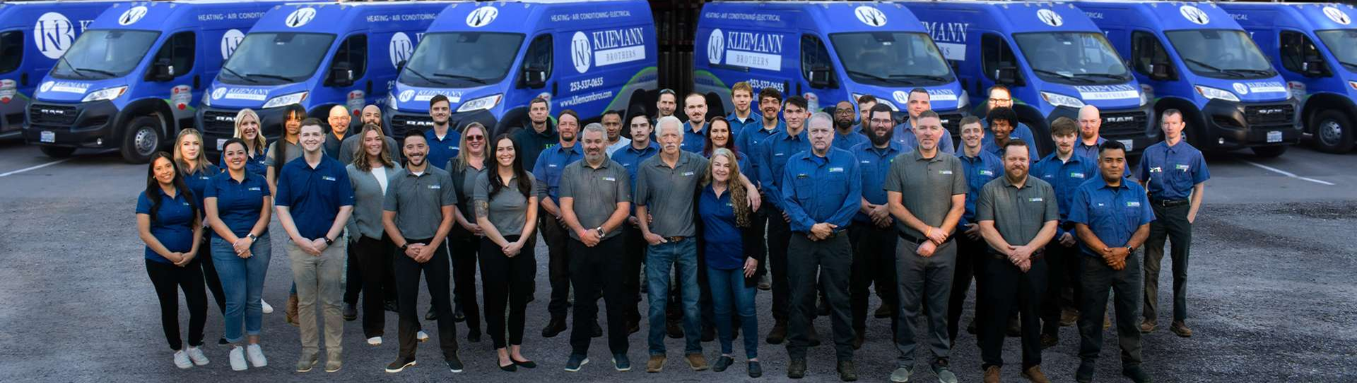 A group of men and women stand in front of a row of blue company vans in a parking lot, all facing the camera and smiling.