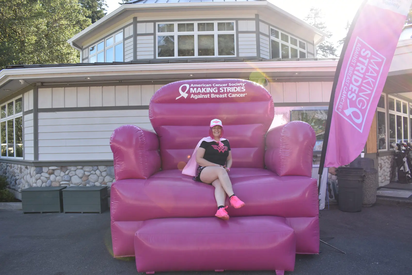 Person sits on an oversized pink chair in front of a house, next to a pink flag promoting Making Strides Against Breast Cancer.