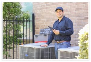 A Kliemann technician in uniform stands outdoors beside HVAC units, holding a tablet and smiling at the camera, ready to assist with your heating needs.