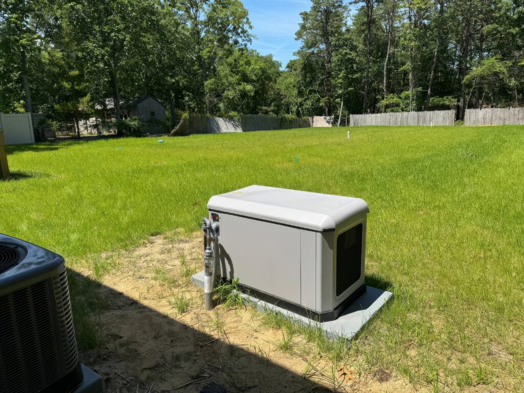 A standby generator is installed on a concrete pad in a grassy backyard near a fence and trees, with utility connections visible.